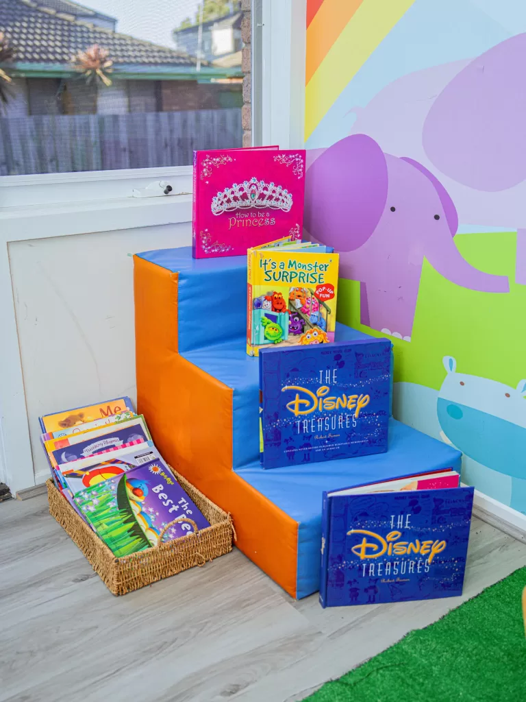 Kinderworld Early Learning Centre classroom reading nook with colourful children's books, soft stairs, and rainbow animal wall mural
